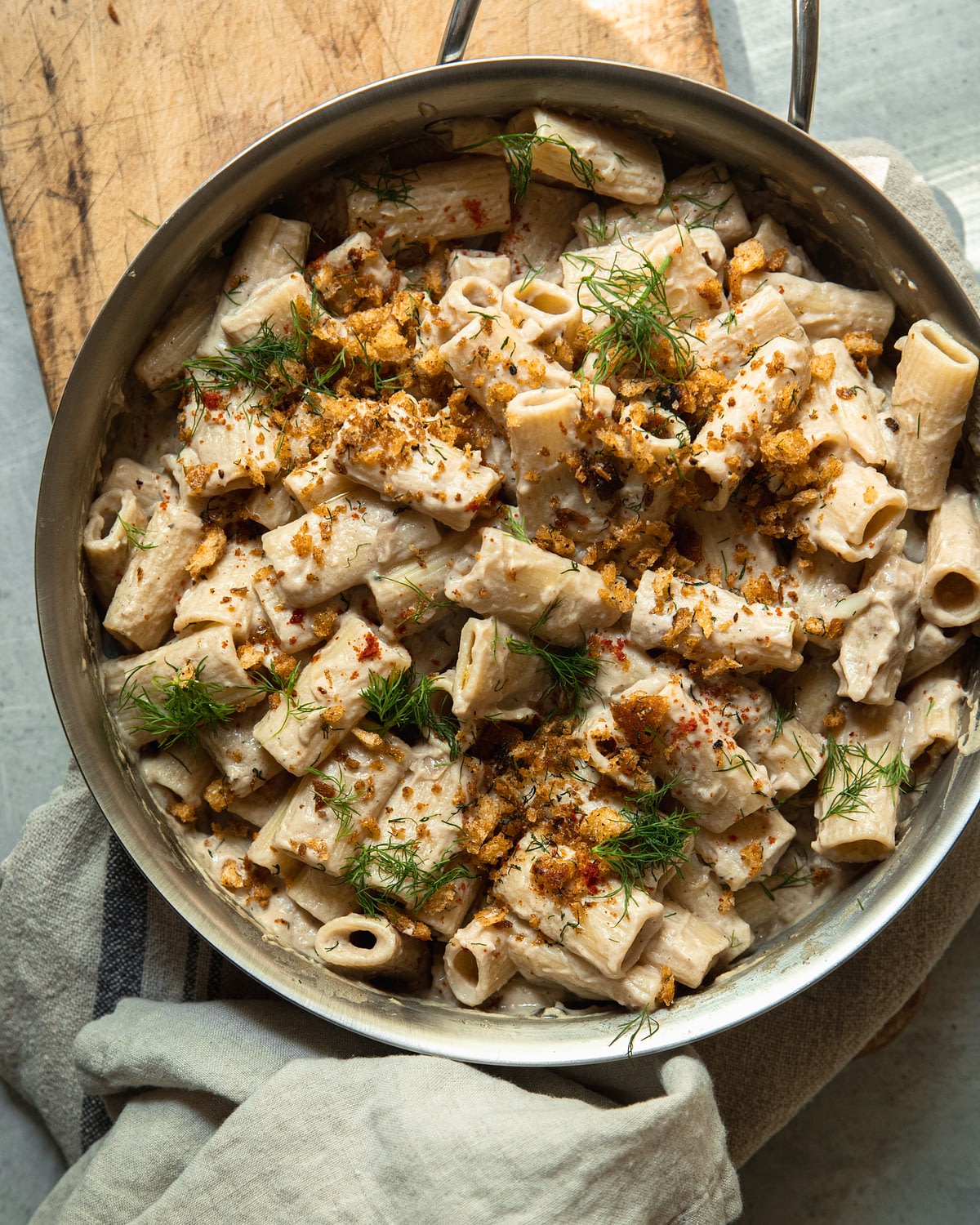An overhead image of a creamy rigatoni pasta in a stainless steel frying pan. The pasta is topped with golden crunchy breadcrumbs, chili flakes, and chopped bits of fresh dill.