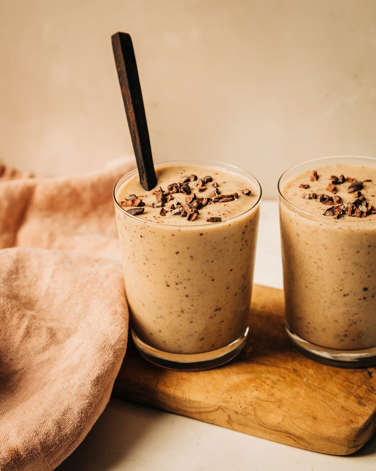 A head on shot of a creamy, beige vanilla chip smoothie in two clear glasses. The smoothie is garnished with cacao nibs and there is a dark wooden spoon sticking out of the one glass.