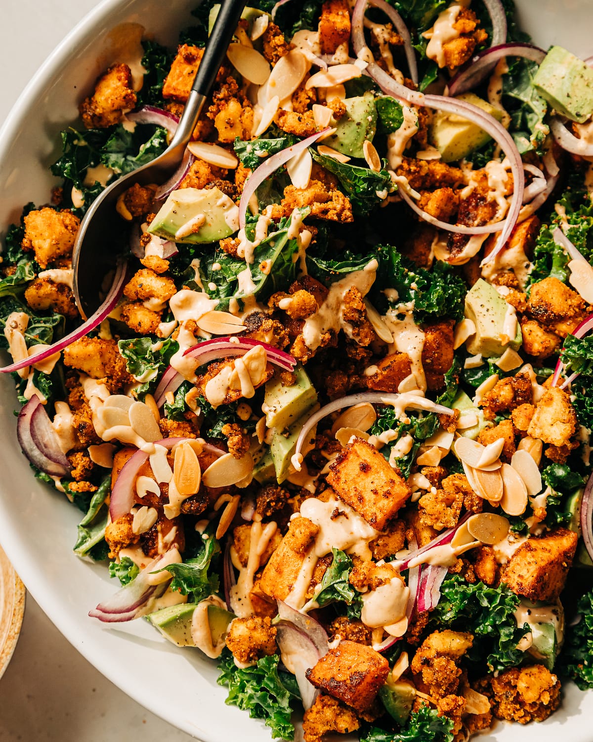 An overhead shot of a kale salad with creamy dressing on top, crispy bits of tofu, chunks of roasted sweet potato, slices of red onion, and cubes of avocado. The salad is in a white bowl.