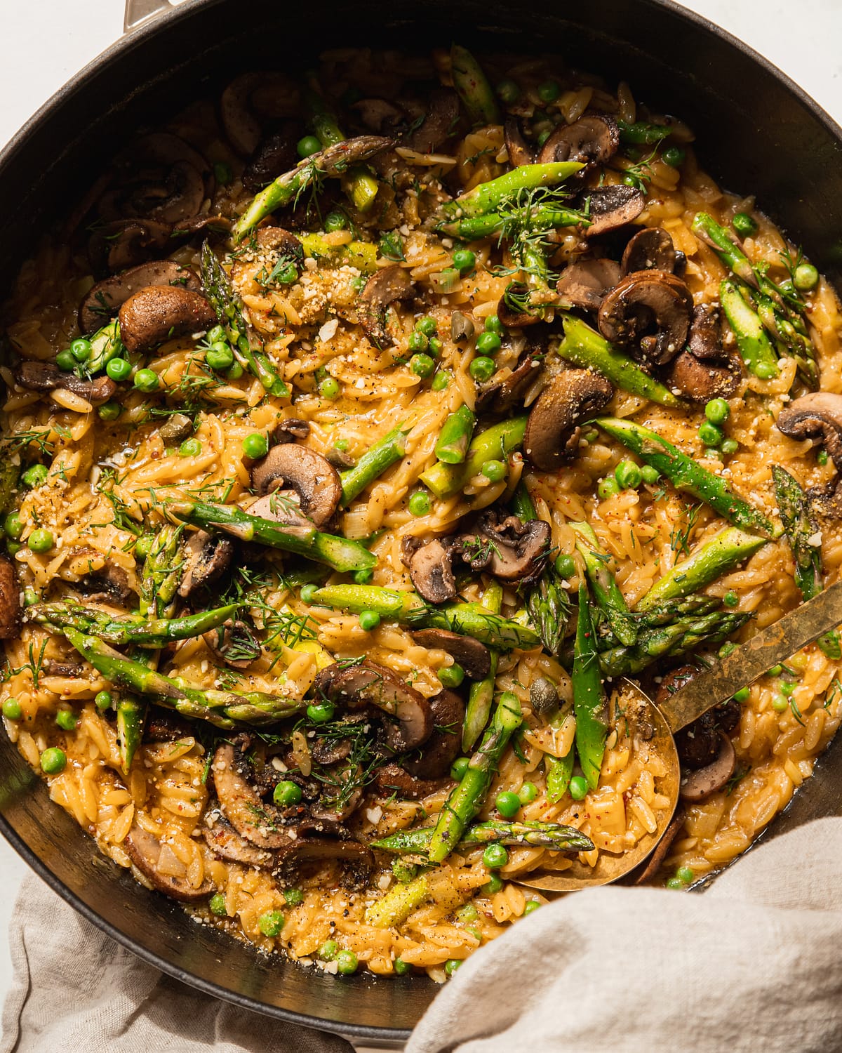 An overhead shot of creamy mushroom asparagus orzo with peas and bits of fresh dill on top. The orzo is in a braiser-style pot with a gold serving utensil sticking out. There is a beige napkin wrapped around the pot handle.