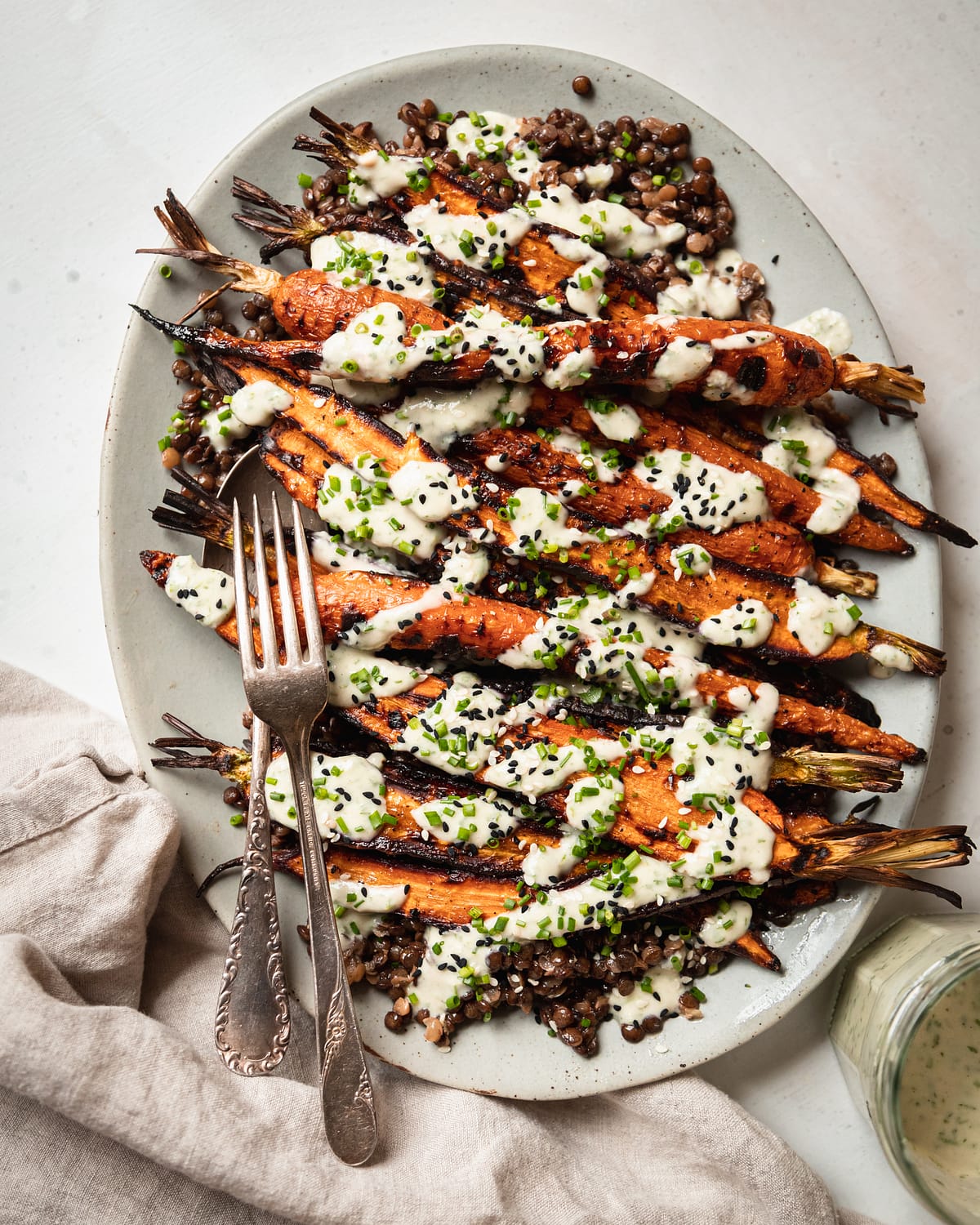 An overhead shot shows a light blue platter topped with cooked french lentils and grilled whole carrots, all topped with a creamy chive-flecked sauce. Extra chives and sesame seeds garnish the dish.