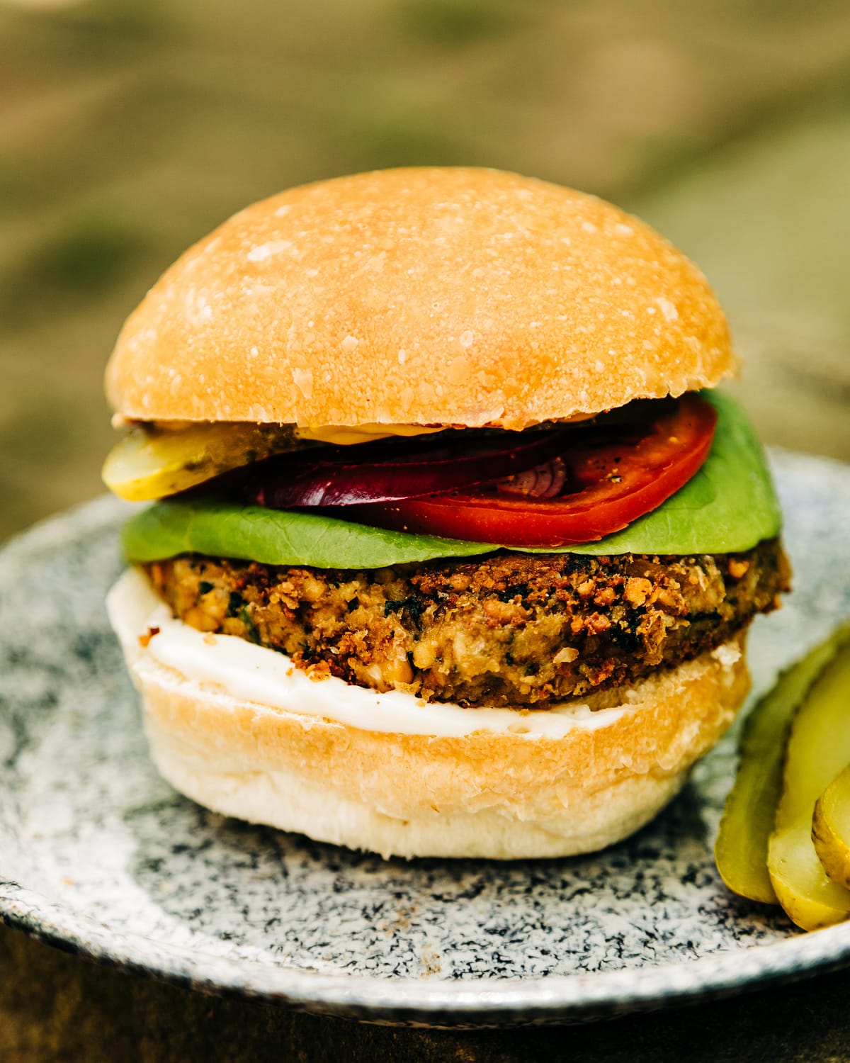 A head on shot of a vegan chickpea umami burger in a bun with lettuce, tomato, pickle, red onion, and mayonnaise as toppings.