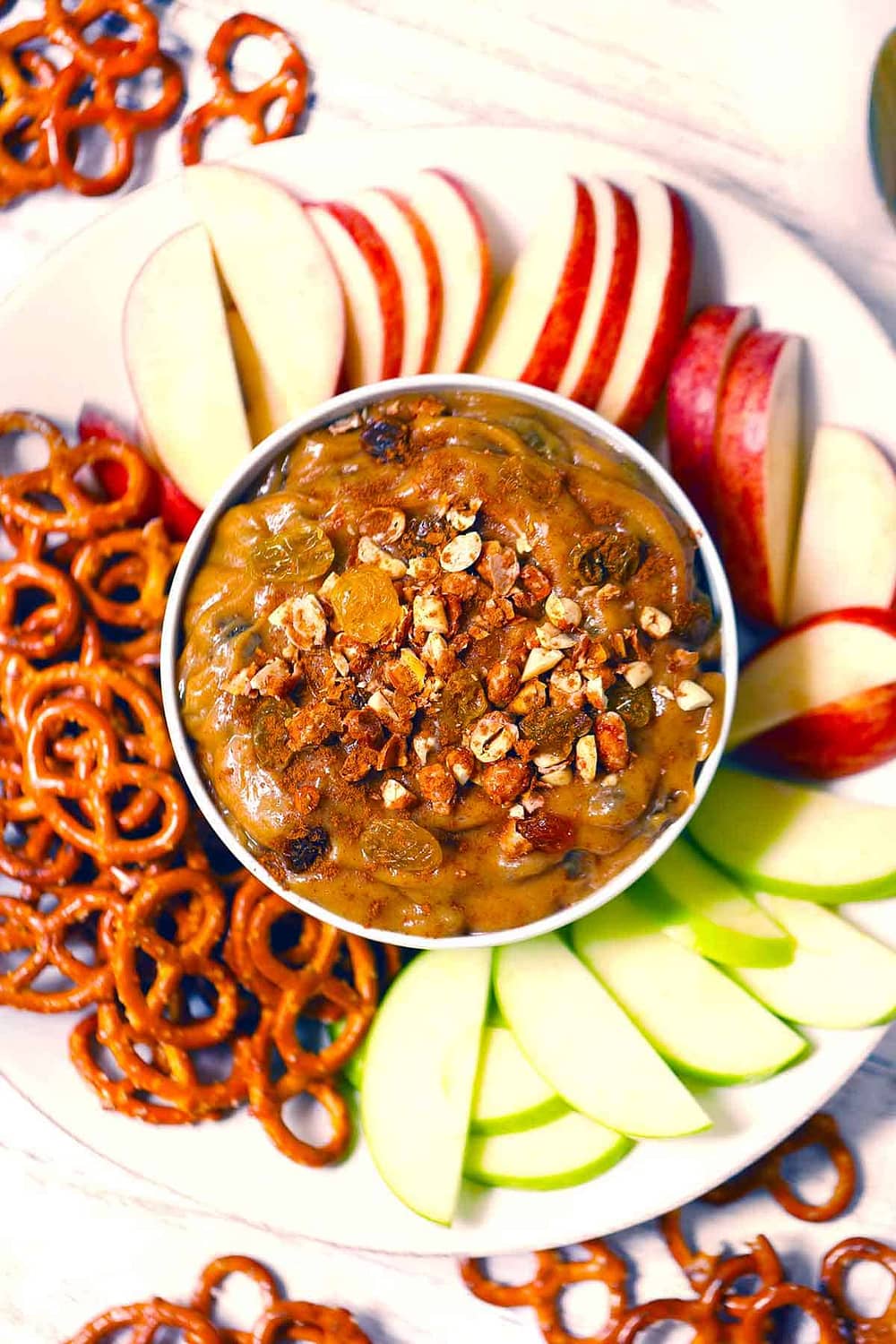Overhead photo of peanut butter dip with raisins on a plate with apple slices and pretzels around it.