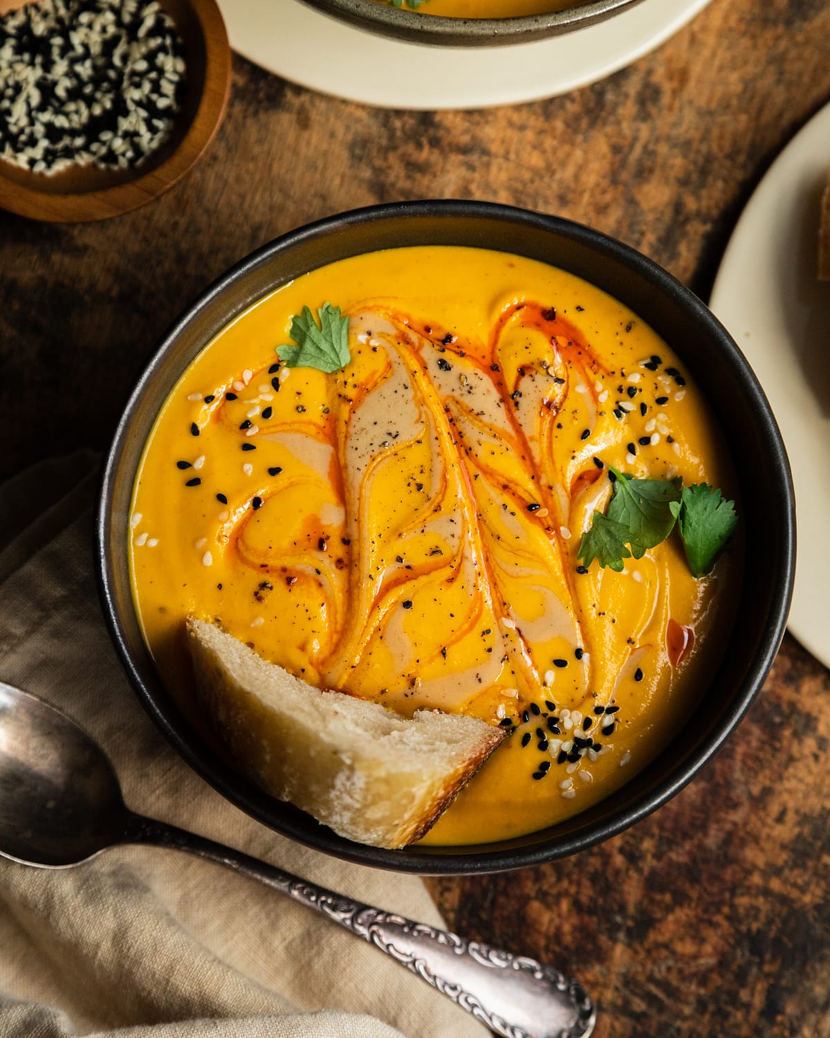 An overhead shot of a bowl of vibrant and creamy orange soup that is swirled with a red slick of oil on top. The soup has a crust of bread in it and is also garnished with sesame seeds and cilantro.