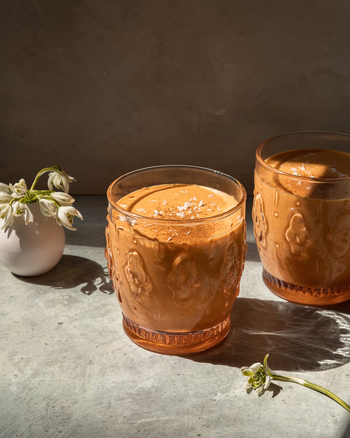 A head on shot of a light orange spring thaw smoothie in two glasses with a small vase of snowdrop flowers in the background.