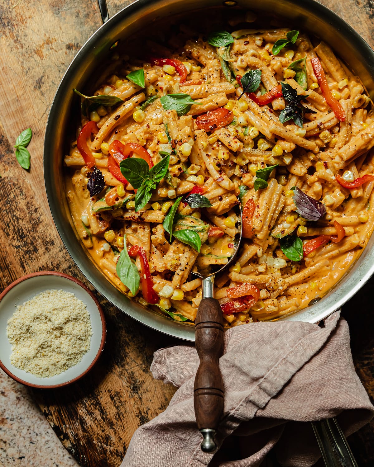 An overhead shot shows a creamy vegan red pepper pasta in a stainless steel pan. The short-shaped pasta is garnished with fresh basil leaves, strips of roasted red pepper, charred corn kernels, dried chili flakes, and freshly ground pepper.