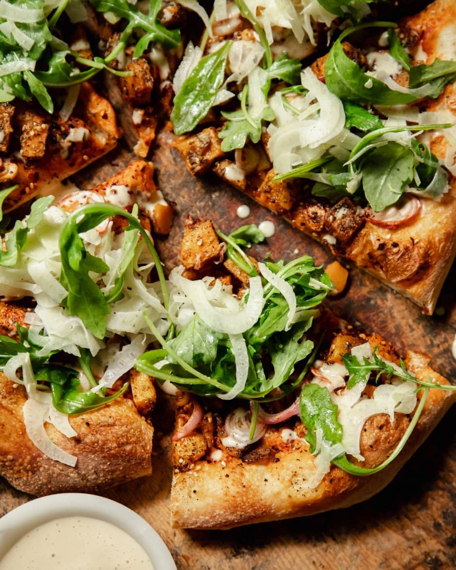 An up close, overhead shot of a round, vegan butternut squash flatbread topped with an arugula fennel salad and drizzles of a creamy white sauce.